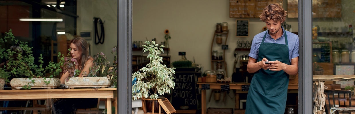 Young entrepreneur with an apron watching his phone in front of his shop